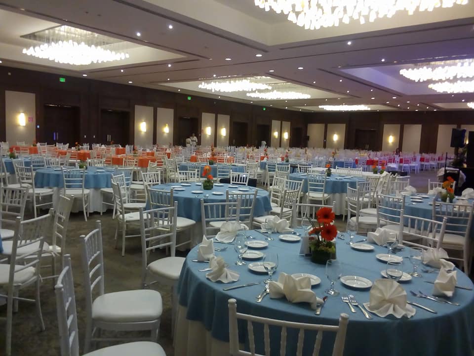 Grand ballroom setup for a MICE banquet at Fiesta Americana Mérida: A wide-angle view of a large, elegant hotel ballroom prepared for a corporate dinner or wedding gala. The room features multiple round tables with light blue tablecloths, white Chiavari chairs, elegant lighting fixtures, and high capacity for convention closing events